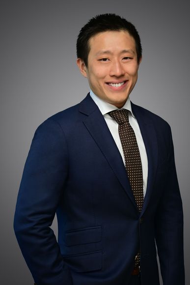 Asian man in a navy suit and patterned tie smiling, against a gray backdrop.