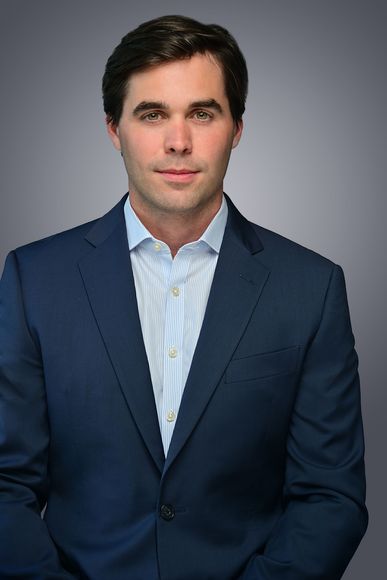 Man in blue suit and striped shirt, smiling slightly, against a gray gradient background.