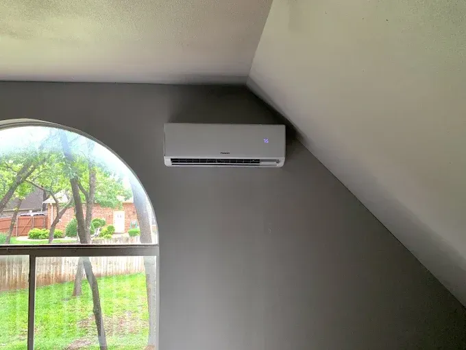 Gray air conditioner mounted on a gray angled wall near an arched window with a view of greenery.