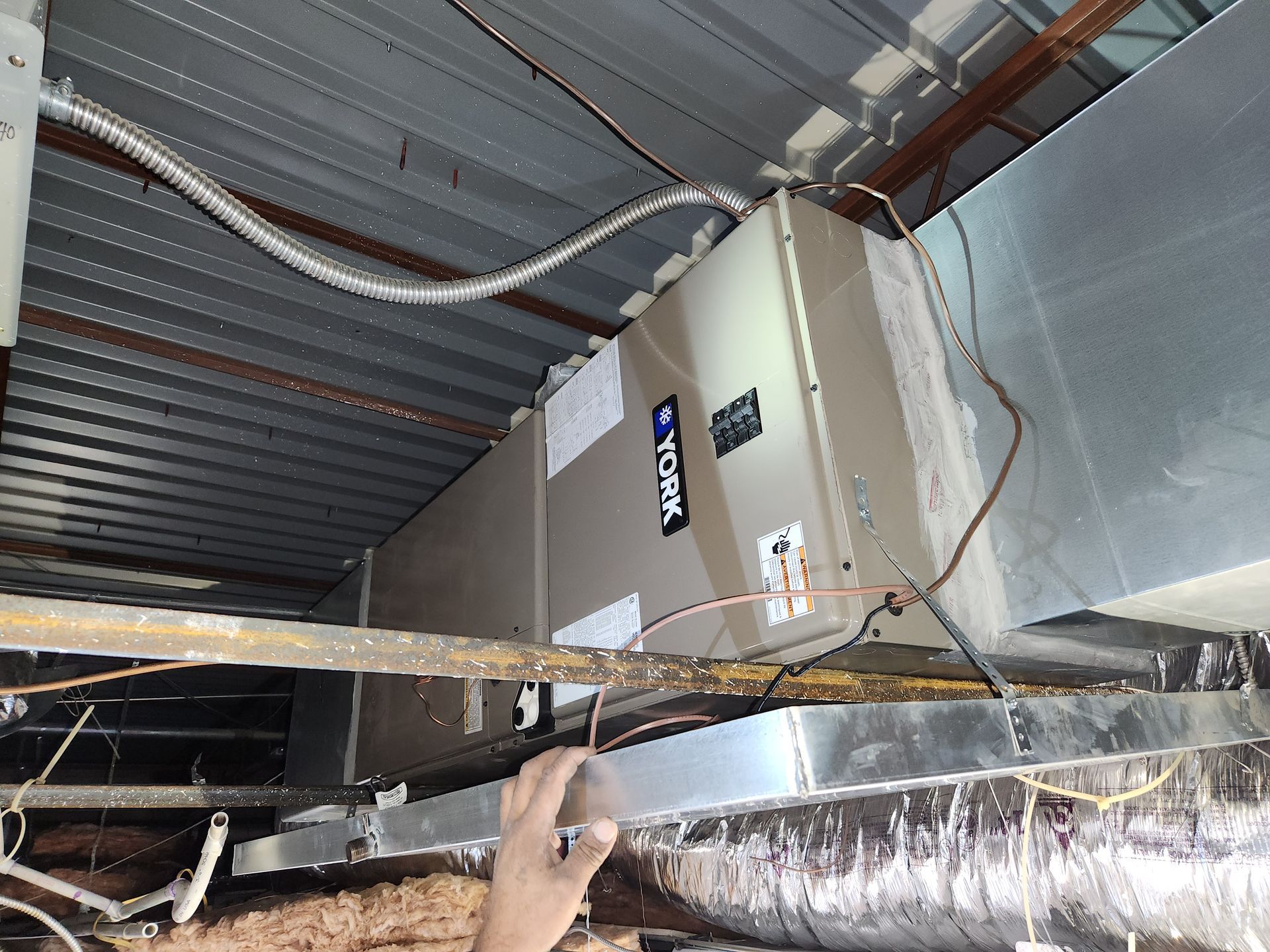 HVAC unit installed in a ceiling cavity, with ductwork and wiring visible. A hand reaches into the frame.