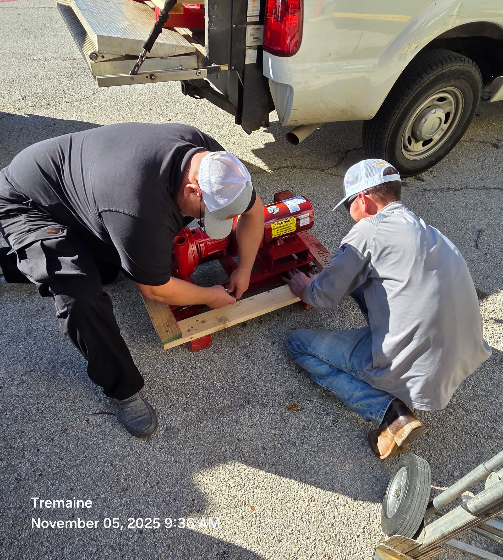 Two workers load red machinery onto a truck. One kneels, the other stands, both wearing hats.