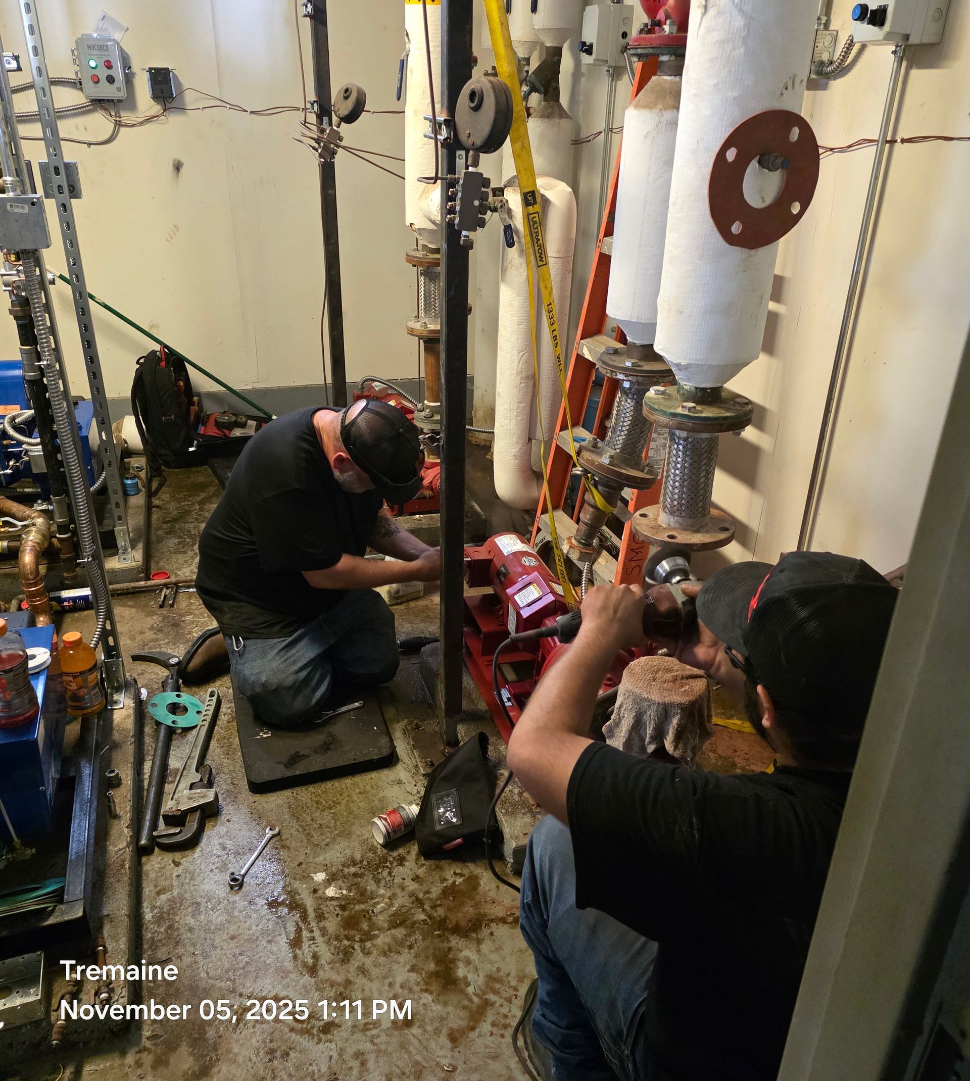 Two workers are repairing machinery in a mechanical room, using tools.