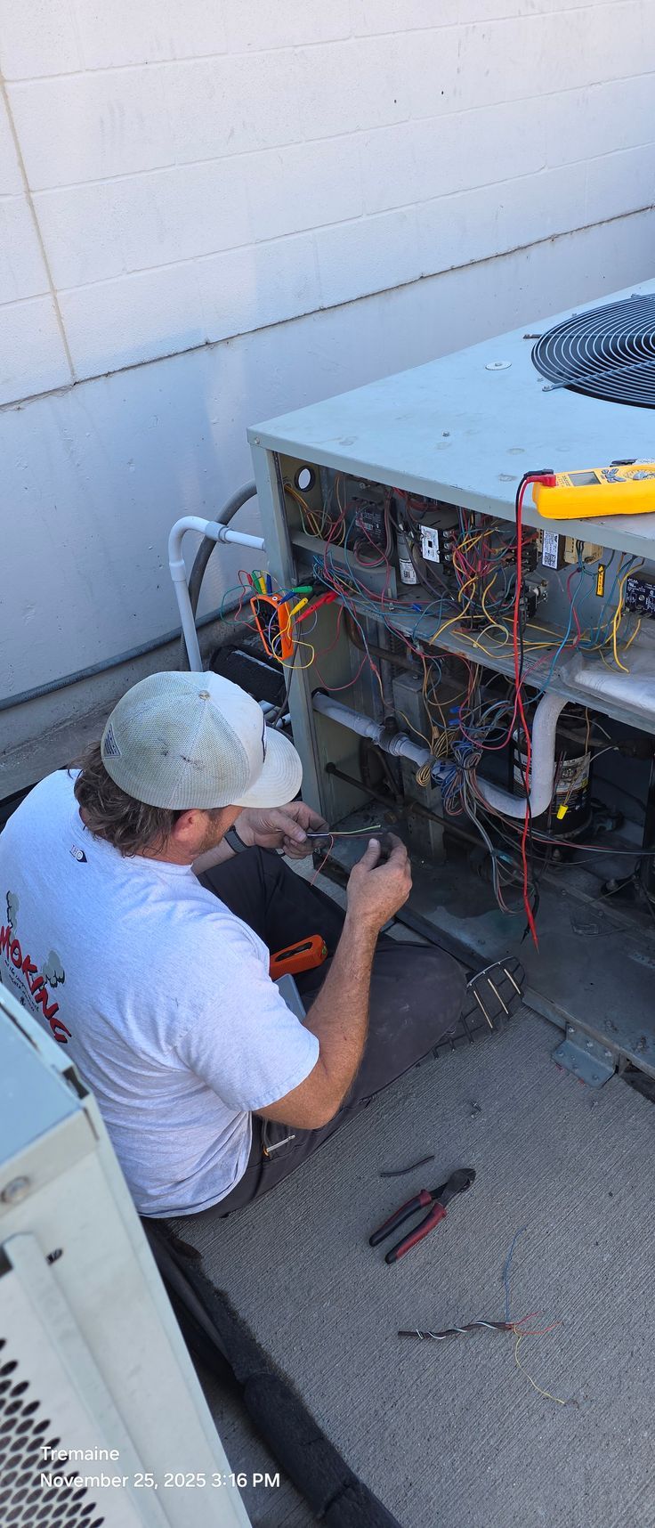 A person works on HVAC wiring on a rooftop. A yellow multimeter sits on the unit, and tools lay beside them.