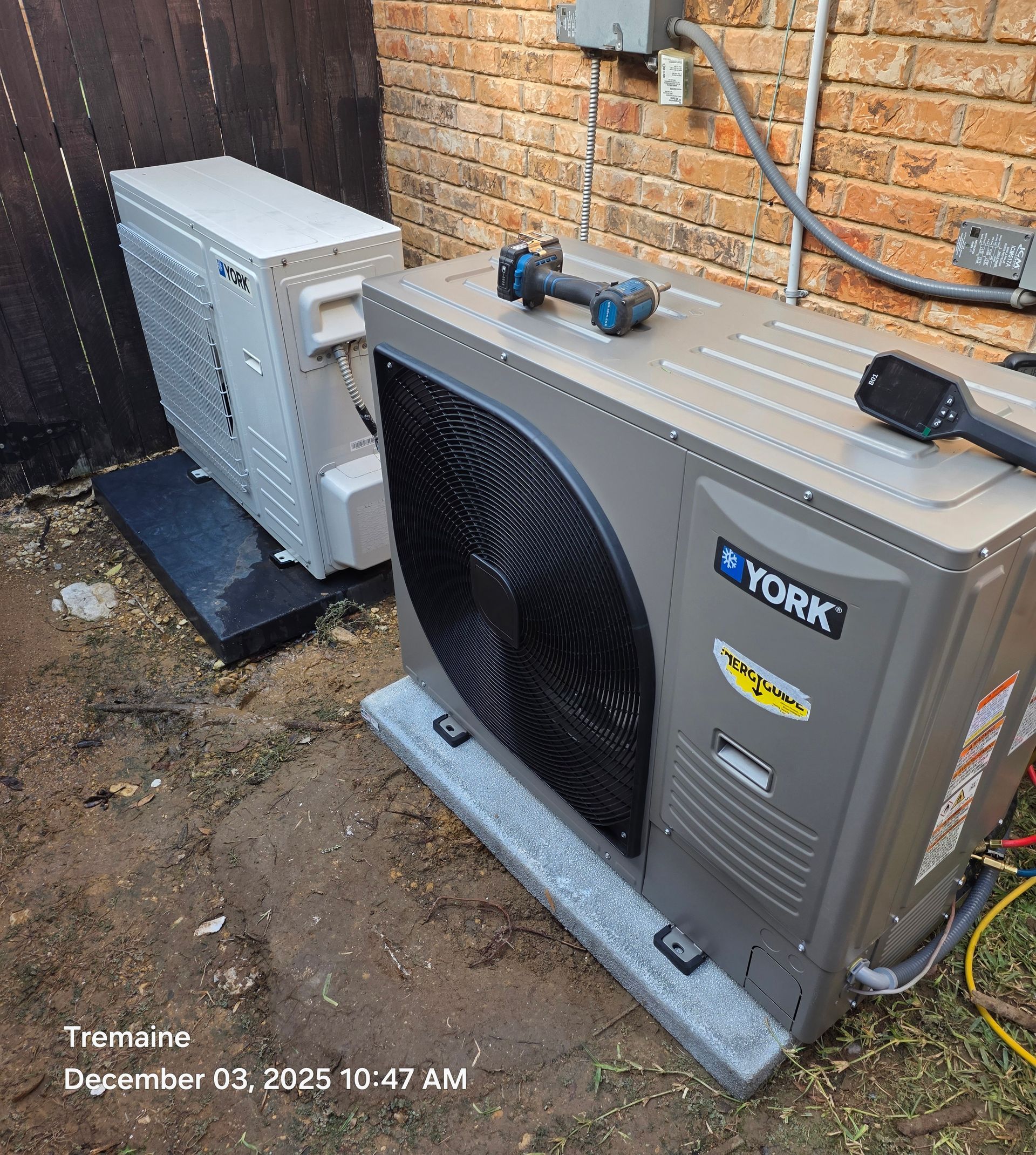 Two HVAC units, one York brand, sit on concrete pads beside a brick wall and fence.