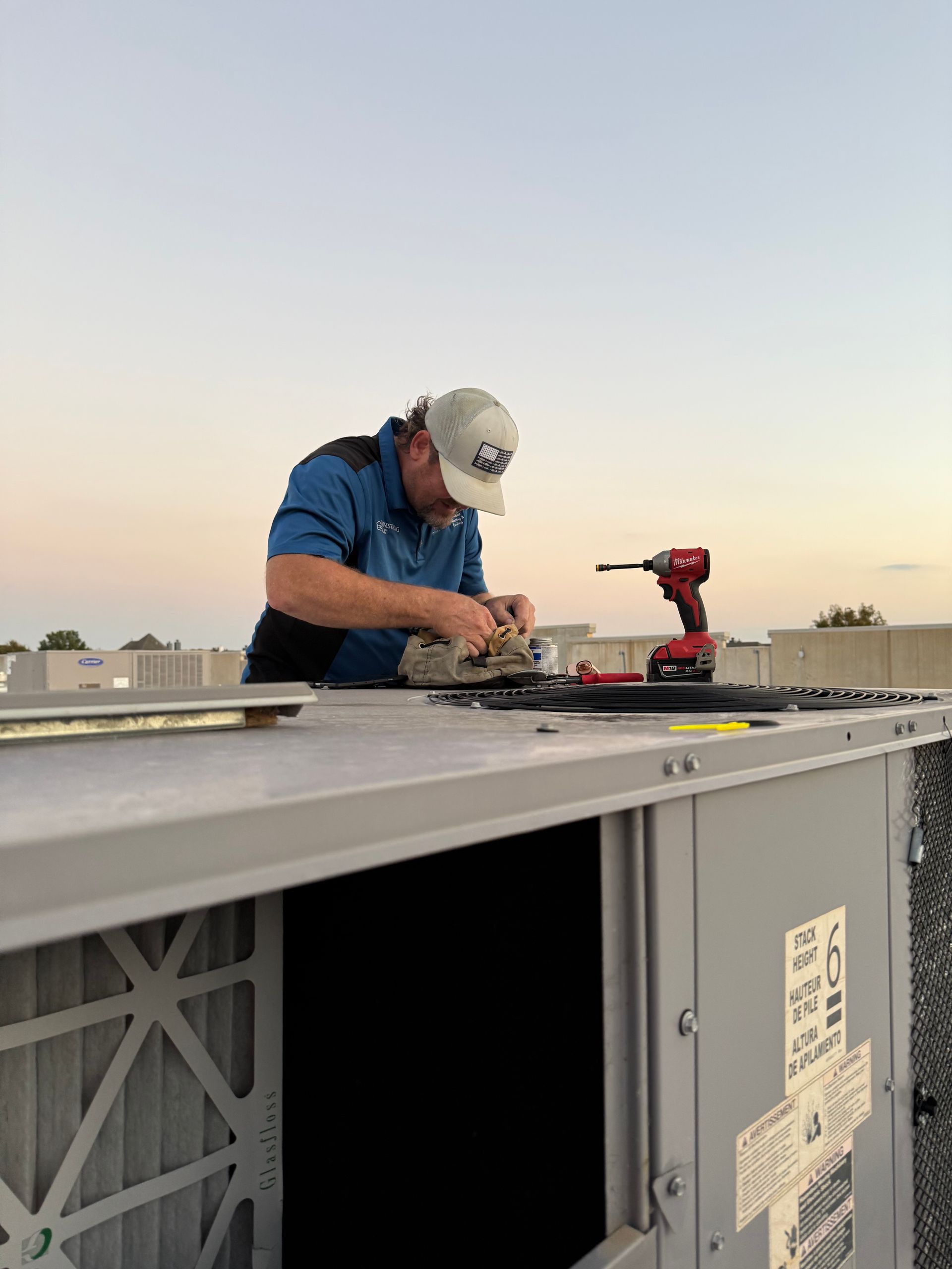 Man in work clothes repairs rooftop HVAC unit with tools at sunset.