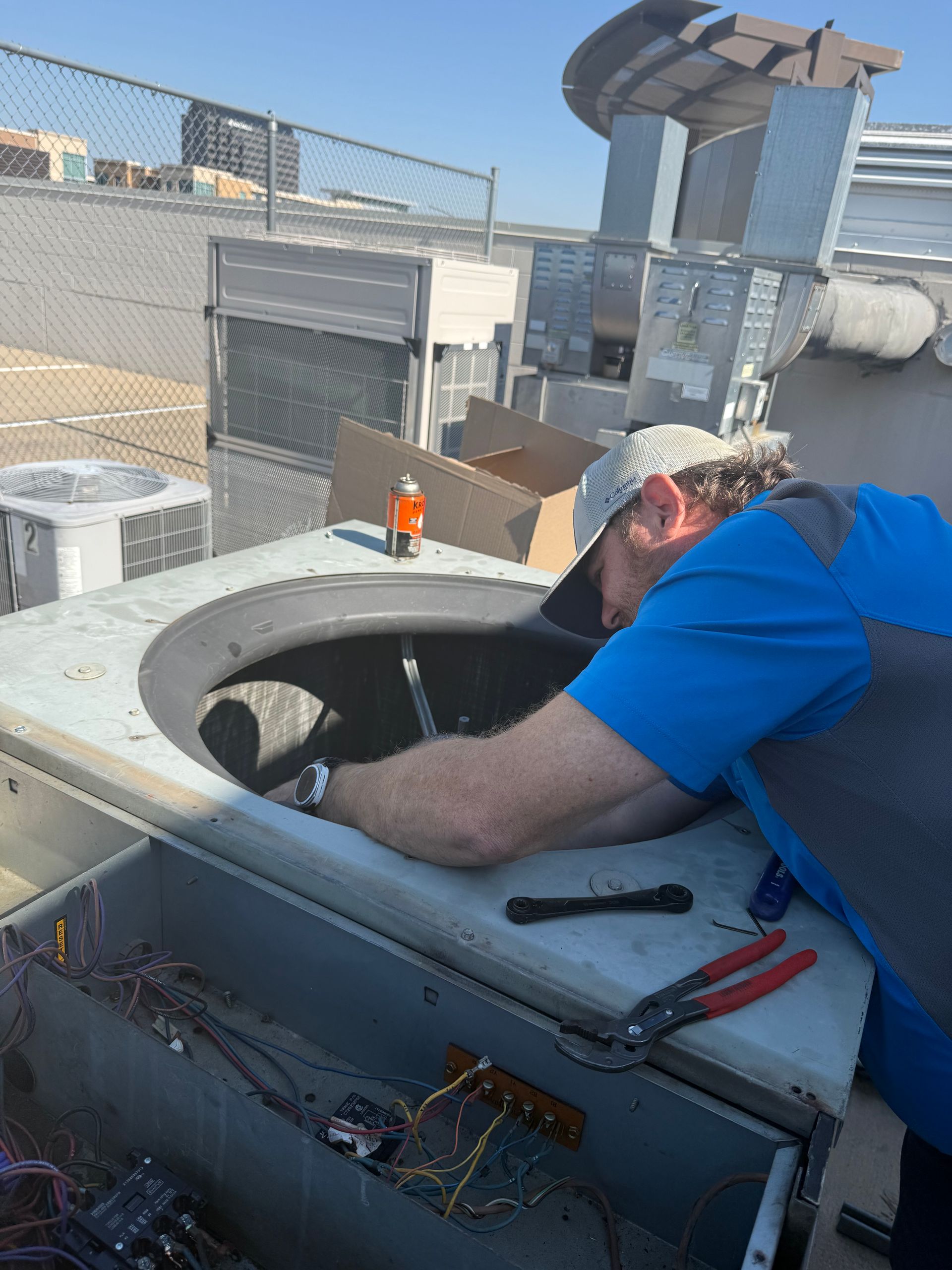 A person in a blue shirt is working on an HVAC unit on a rooftop, with tools visible.