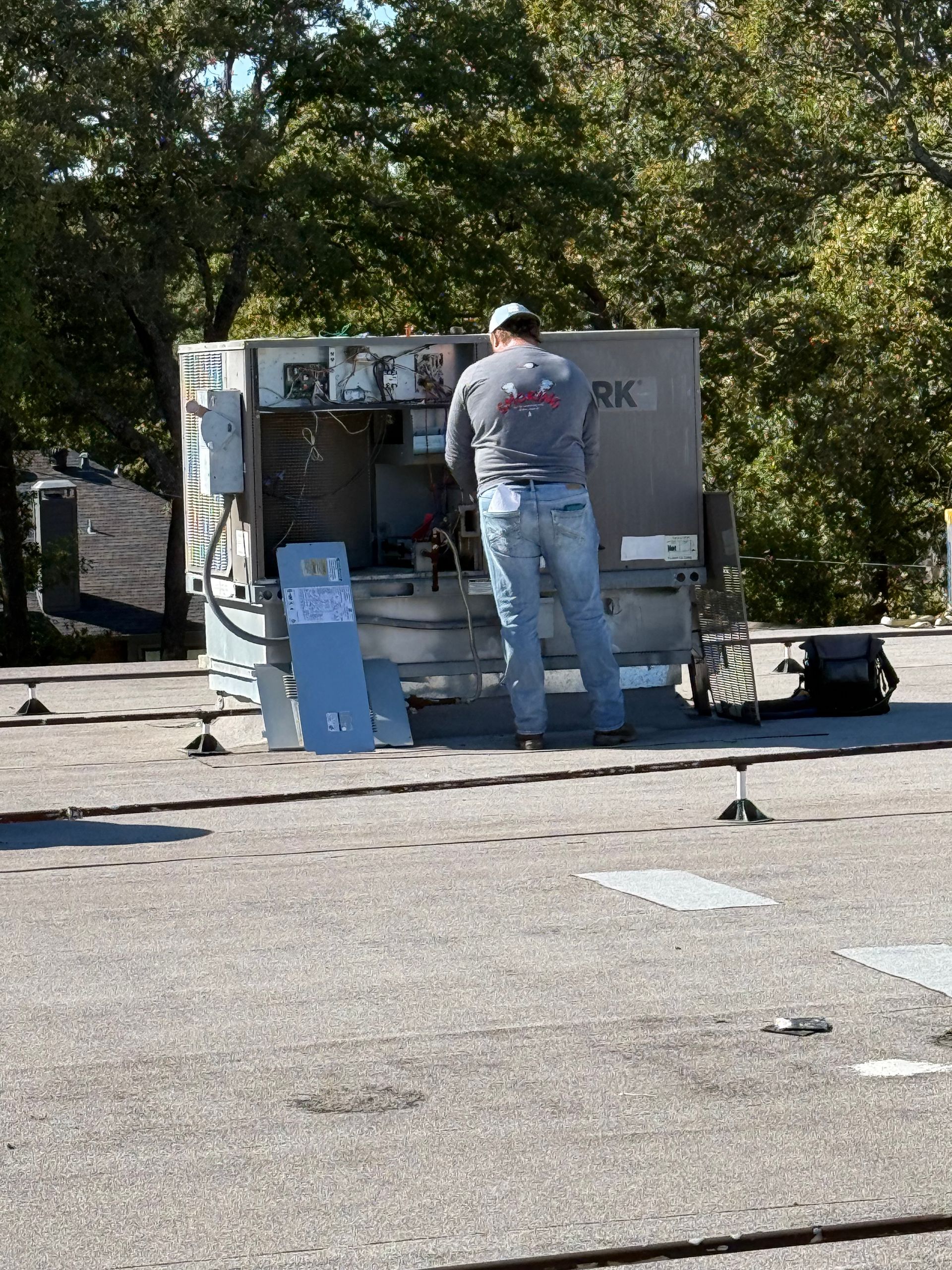 Person working on rooftop HVAC unit. Gray shirt, blue jeans. Outdoors, sunny day.