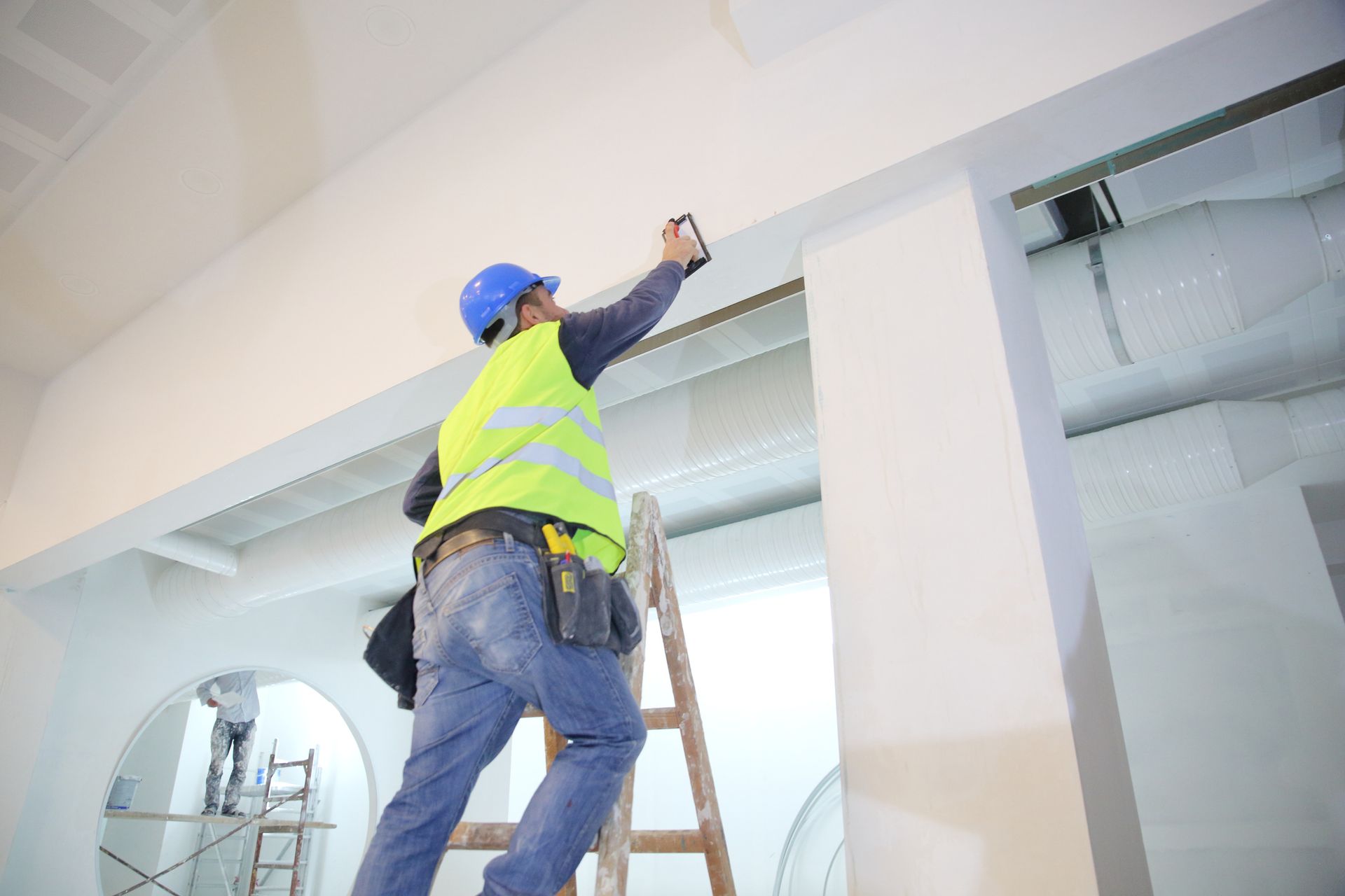 A construction worker is standing on a ladder measuring a wall.