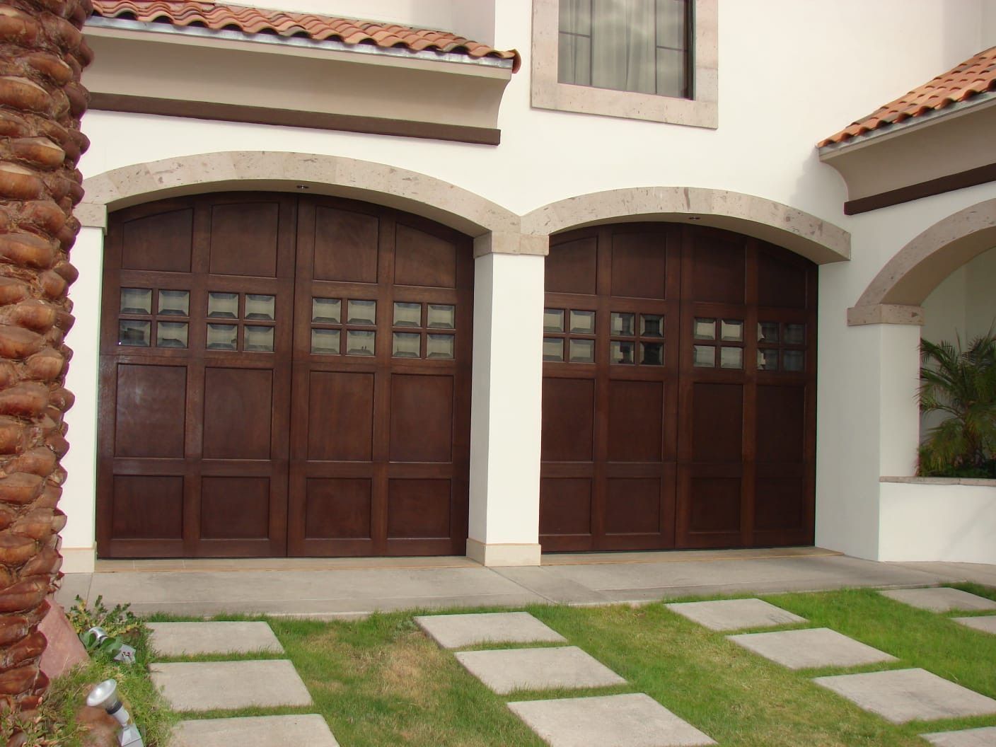 Two dark wooden garage doors with arched stone trim on a white building, with a lawn and walkway.