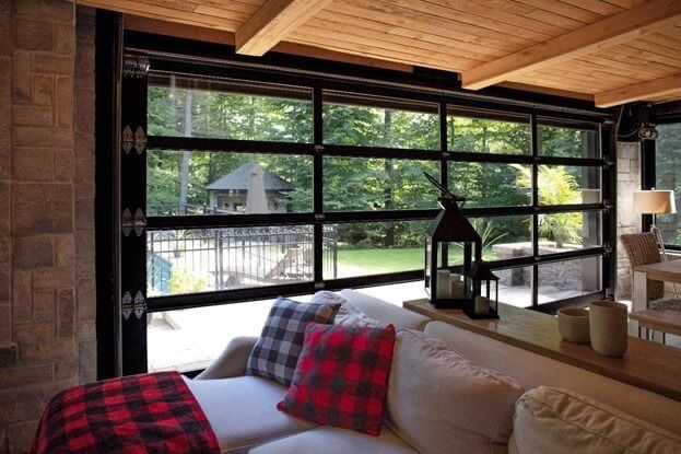 Living room with large glass garage door open to a backyard. Couch with plaid pillows, wooden ceiling, stone wall.