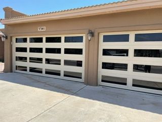 Two beige garage doors with rectangular glass panels, on a stucco house under a blue sky.