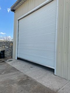 Closed, horizontal-striped, gray overhead door on a tan industrial building. Concrete apron. Blue sky.