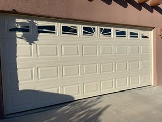 Cream-colored garage door with rectangular and geometric window panel. Shadow of a car is visible.