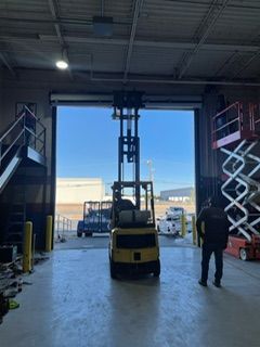 Forklift in warehouse doorway, person standing nearby, lift in raised position, clear sky visible.