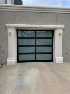 Garage door with black frame and frosted glass panels, flanked by lights and decorative white trim.