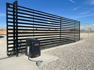 Black metal sliding gate with automatic opener on gravel, blue sky background.
