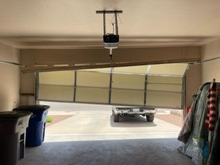 Garage door partially open, bent inward. Car is parked in the driveway. Two trash cans sit inside the garage.