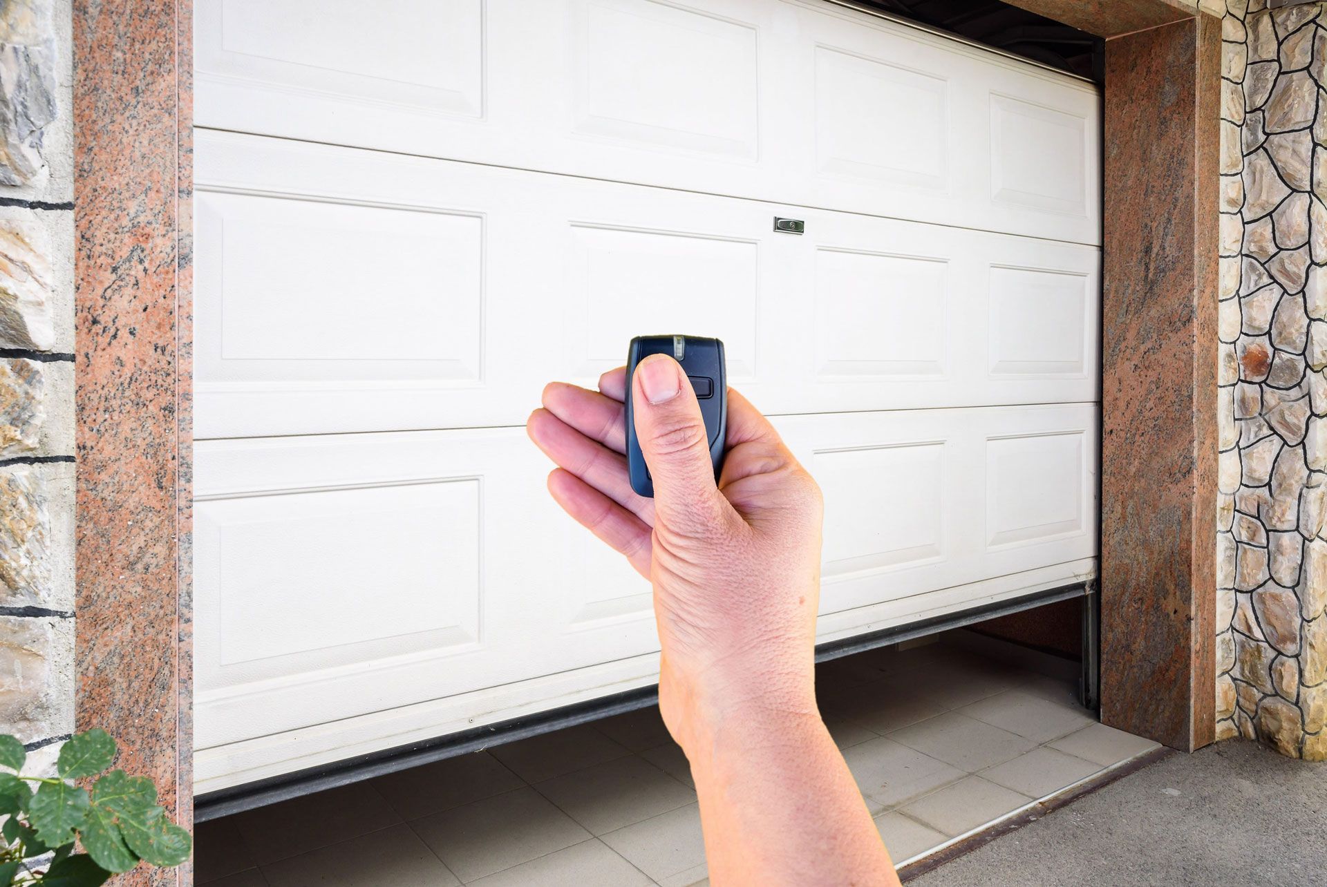 Hand holding a remote opener toward a white garage door in a stone-framed driveway