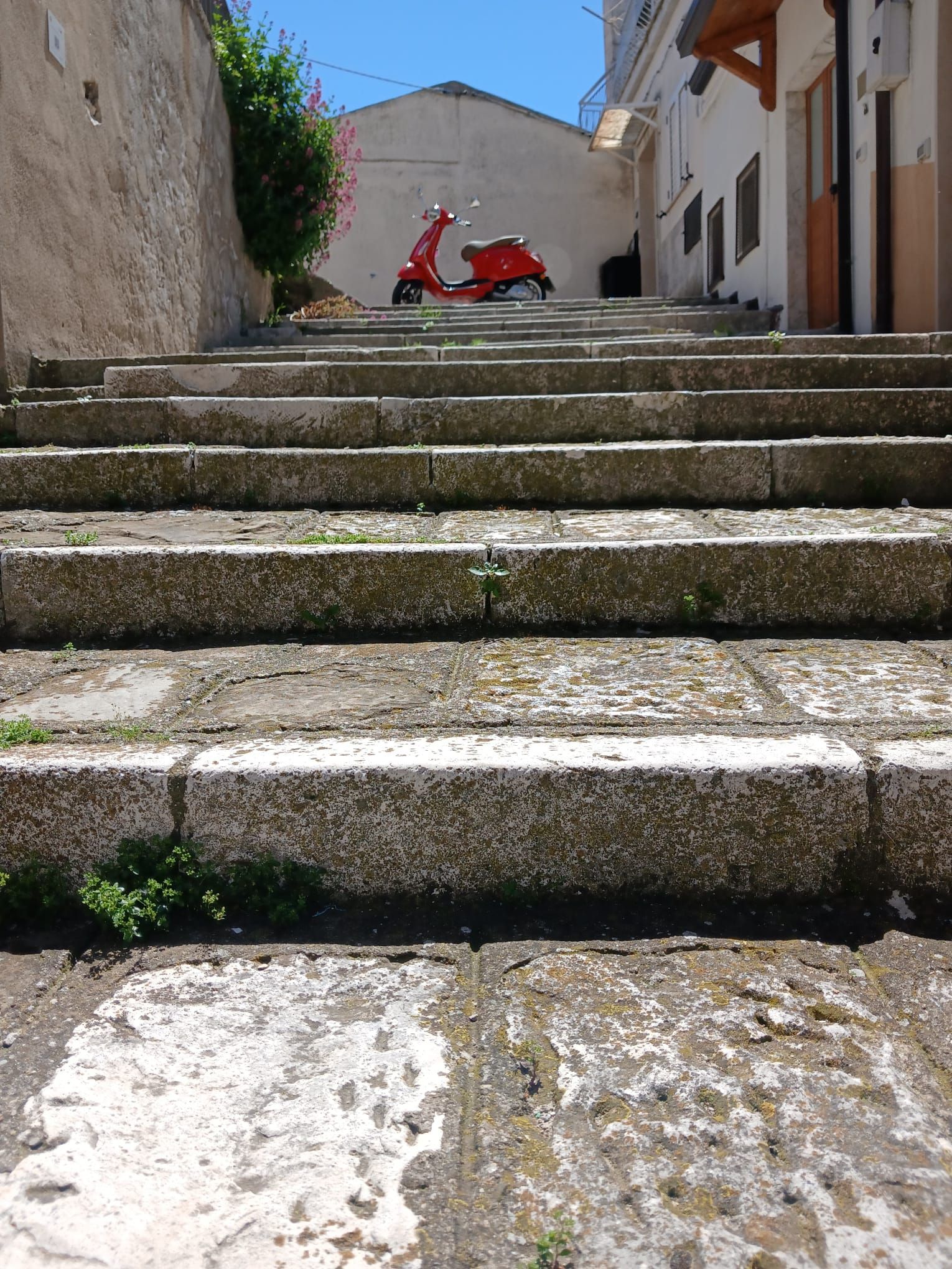 Stone steps leading up to a red scooter in a sunny alley between white buildings.