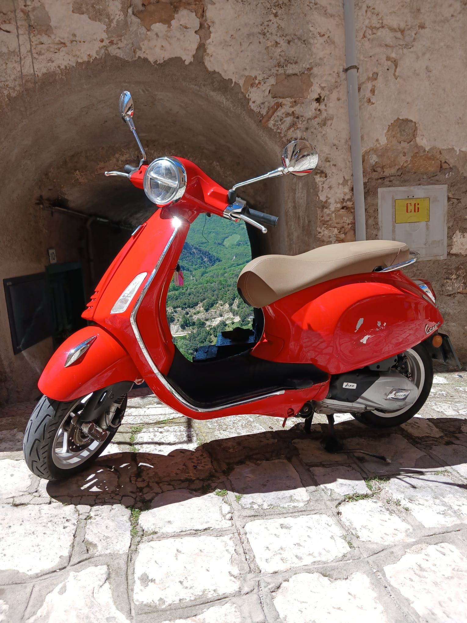 Red scooter parked on a cobblestone path under an archway, with a beige seat and chrome mirrors.