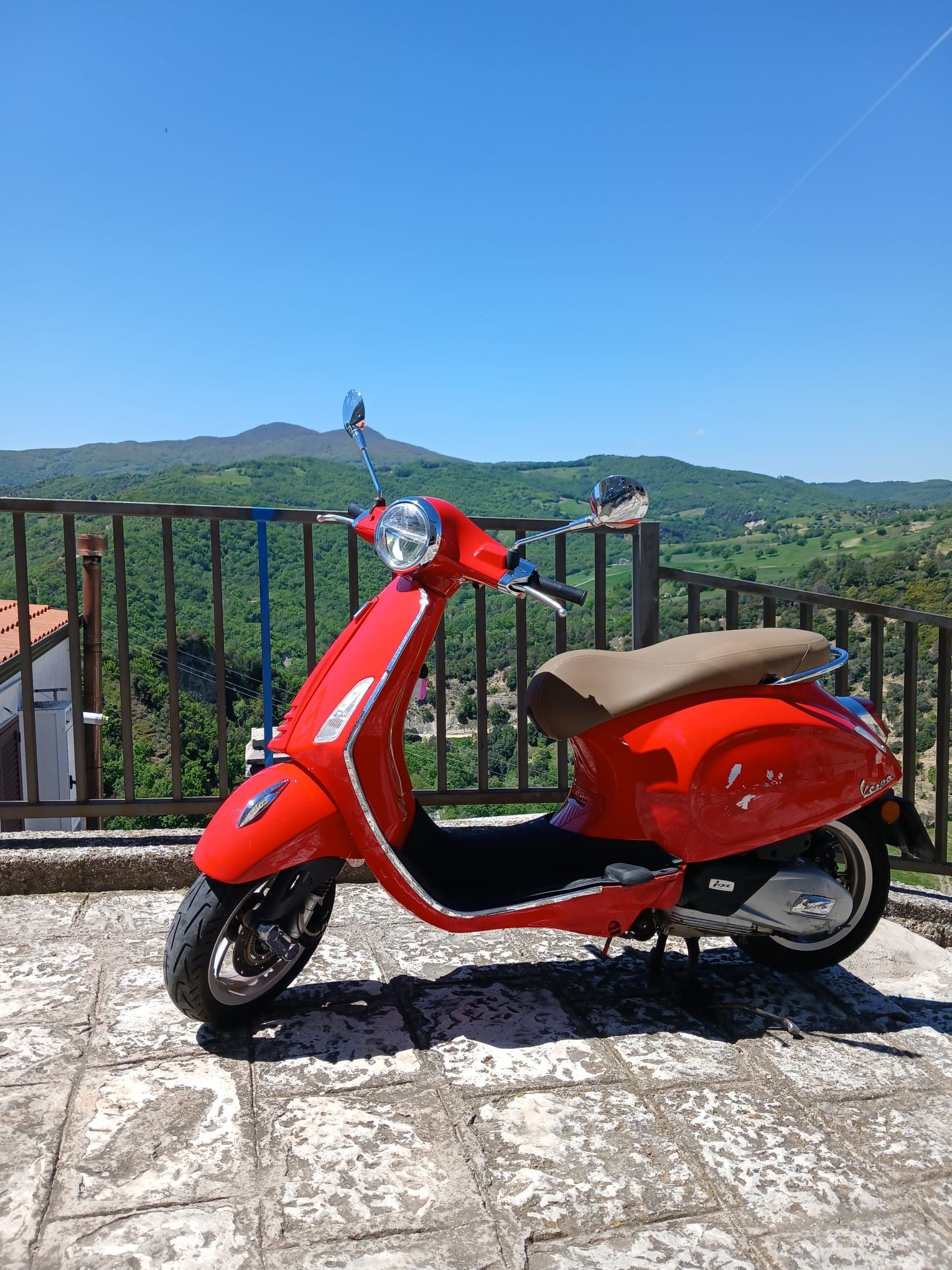 Red scooter parked on a stone terrace overlooking green hills and a blue sky.