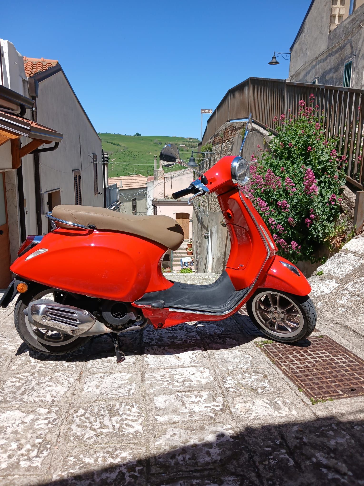 Red scooter parked on a sunlit cobblestone street between houses and flowers.
