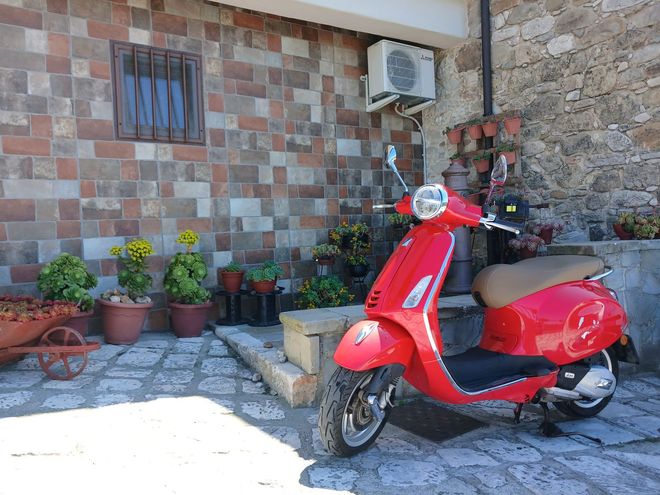 Scooter rosso parcheggiato accanto all'ingresso di una casa in pietra e tegole con piante in vaso e un condizionatore d'aria.