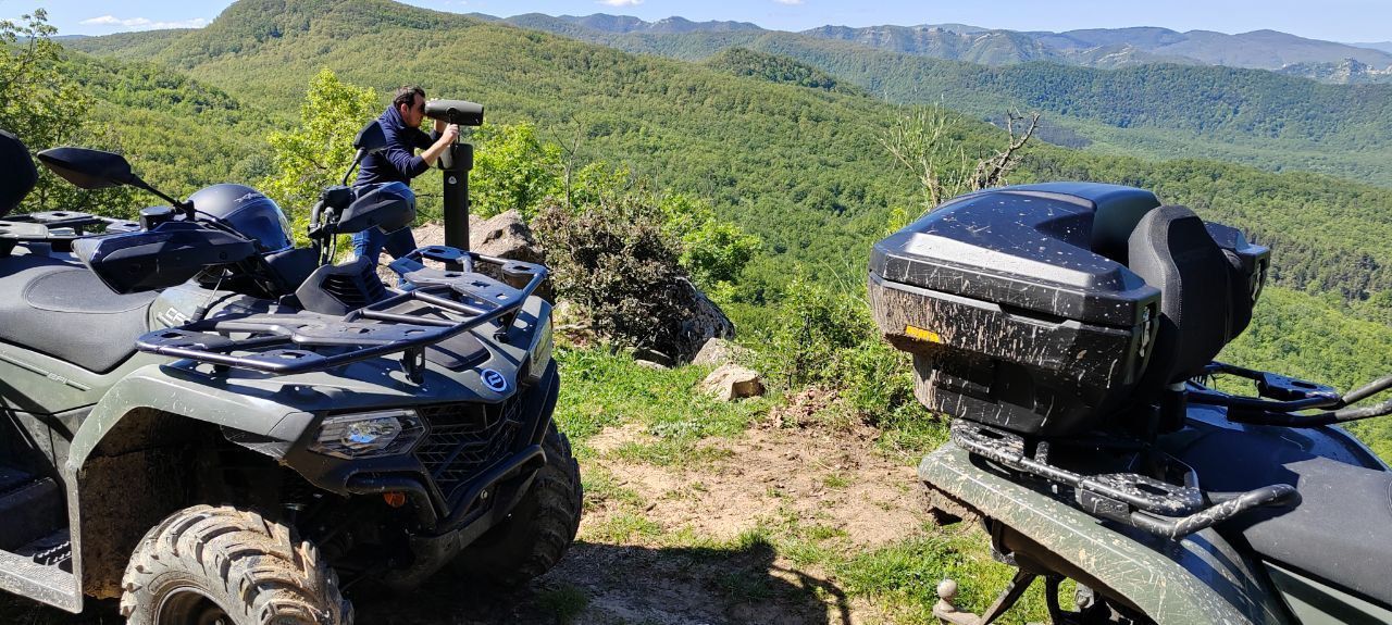 Two ATVs parked on a grassy hillside overlooking a green mountain valley, with a person standing nearby.