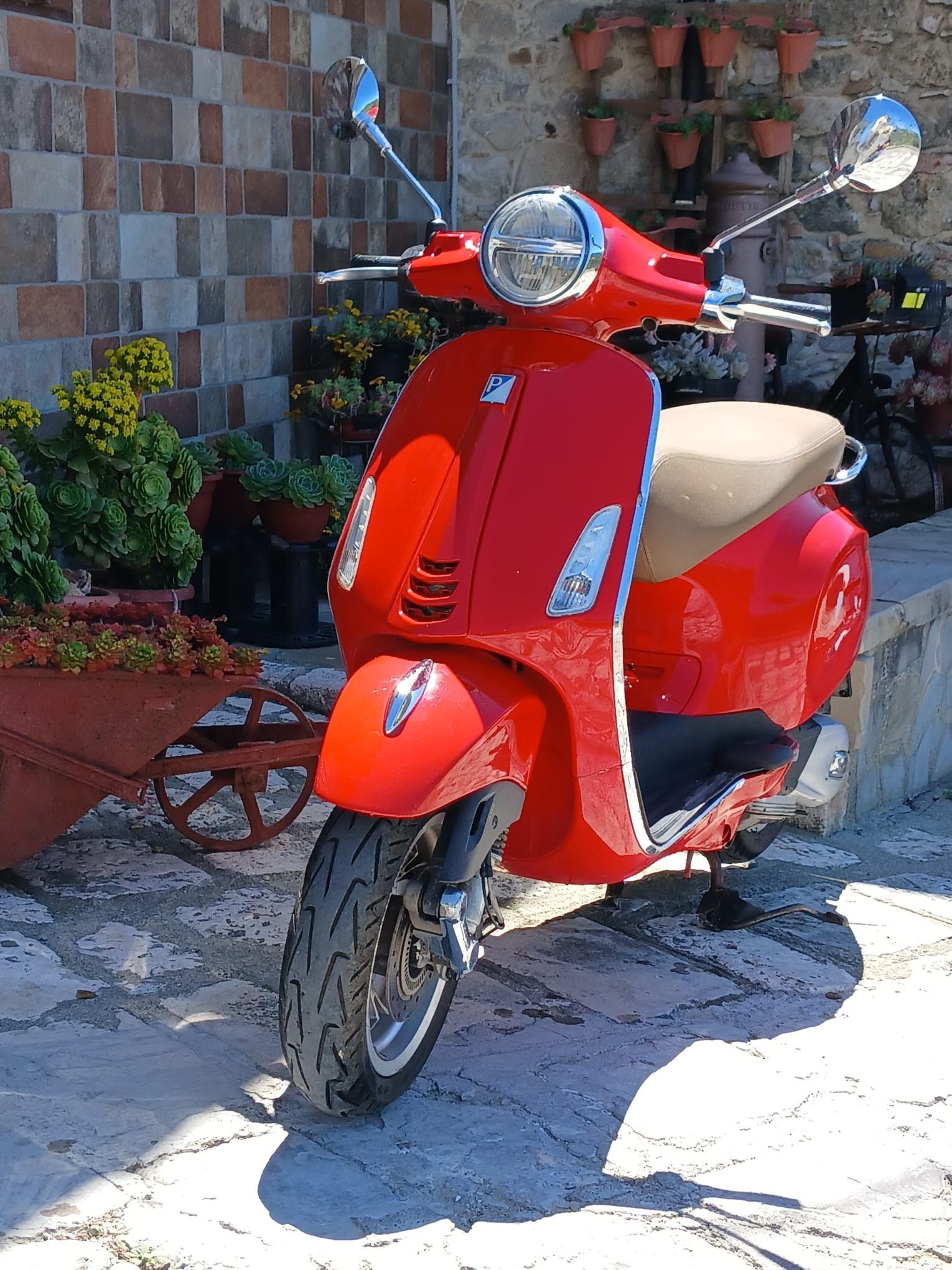 Red scooter parked beside a brick wall in a sunny outdoor courtyard.