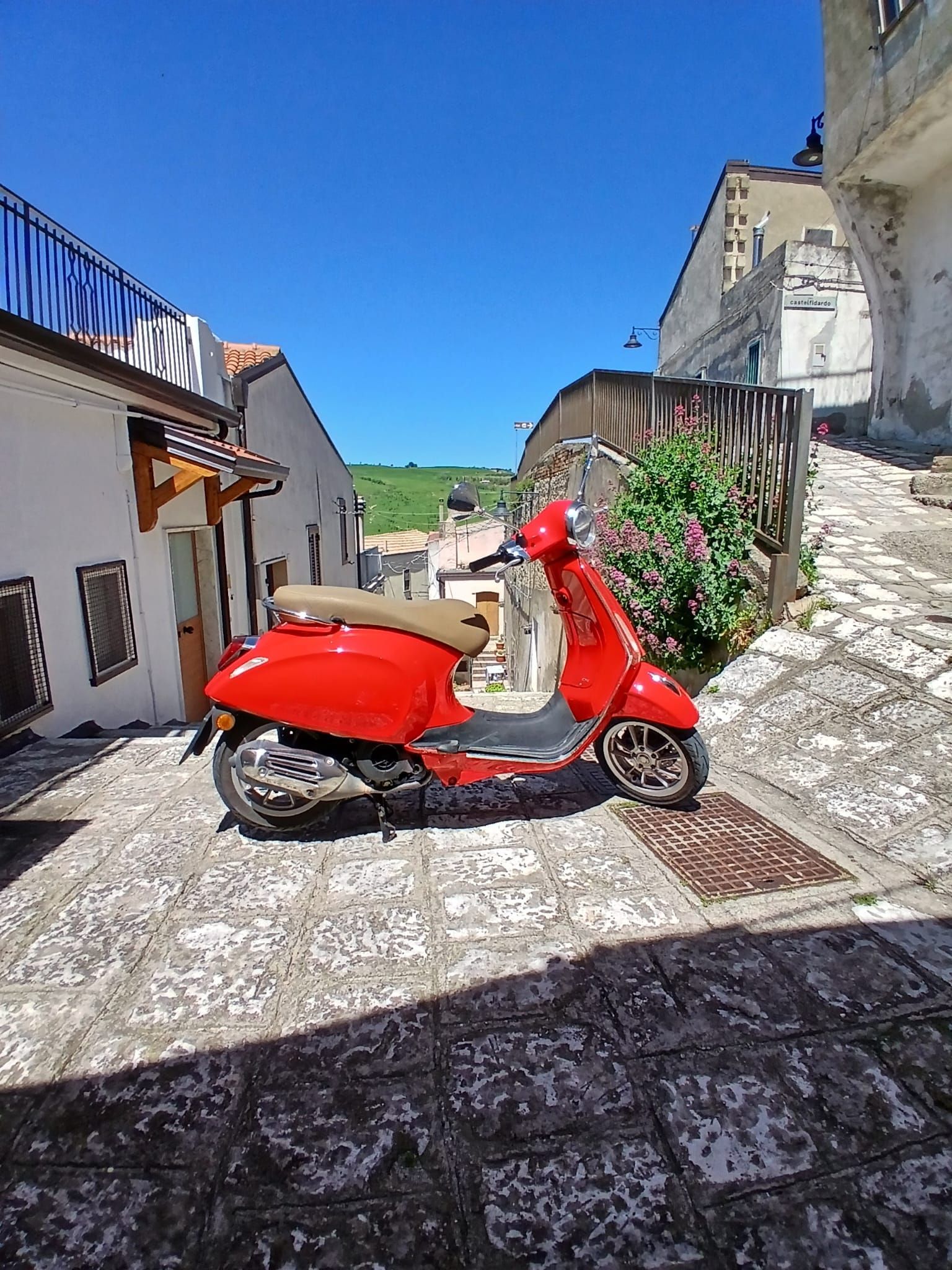 Red scooter parked on a sunlit cobblestone street between white buildings under a clear blue sky