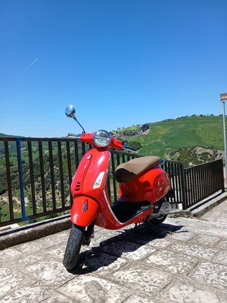 Uno scooter rosso parcheggiato su una terrazza in pietra accanto a una ringhiera nera, con colline verdi e cielo azzurro sullo sfondo.