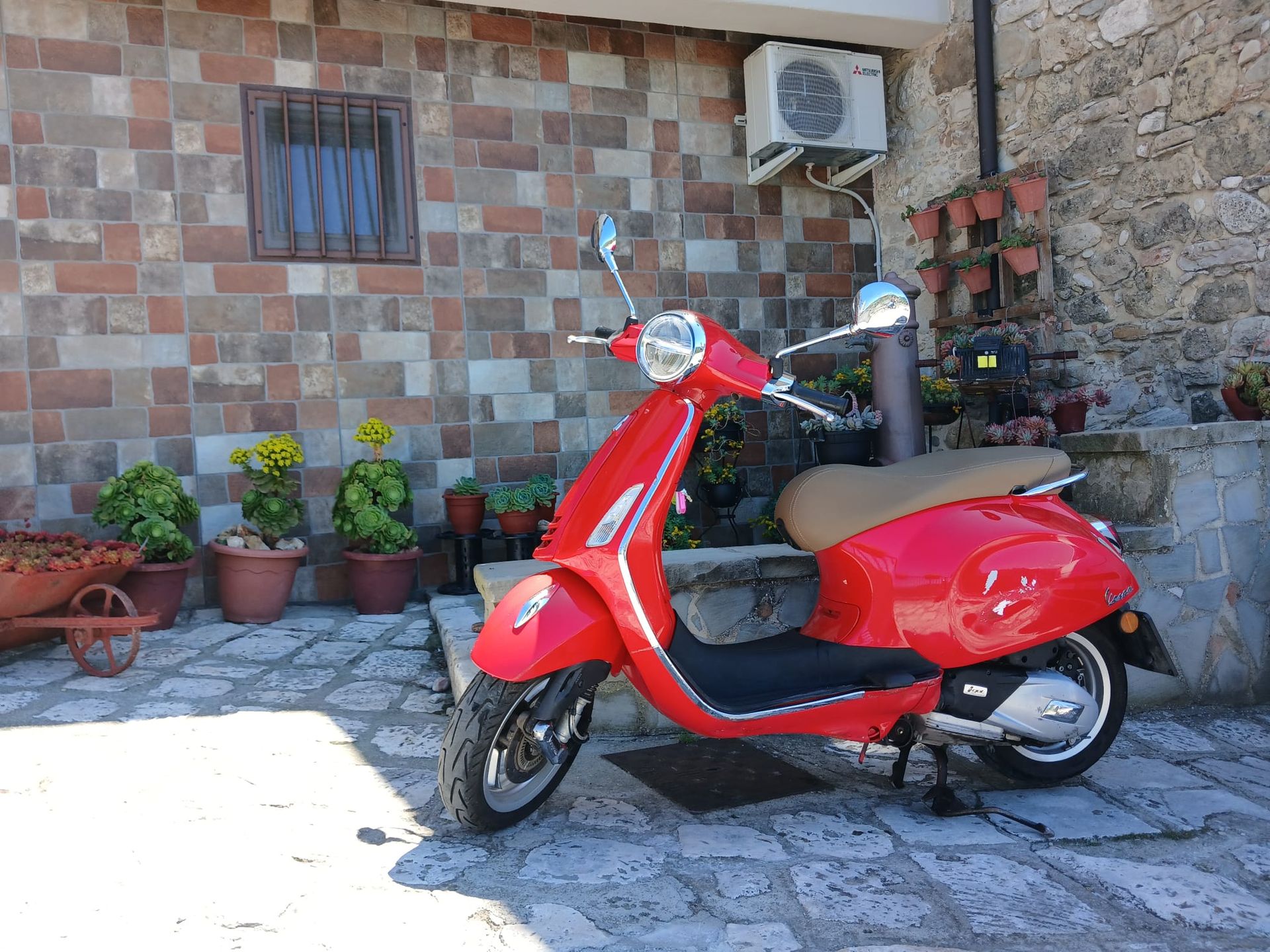 Red scooter parked on a stone patio beside potted plants and a tiled wall