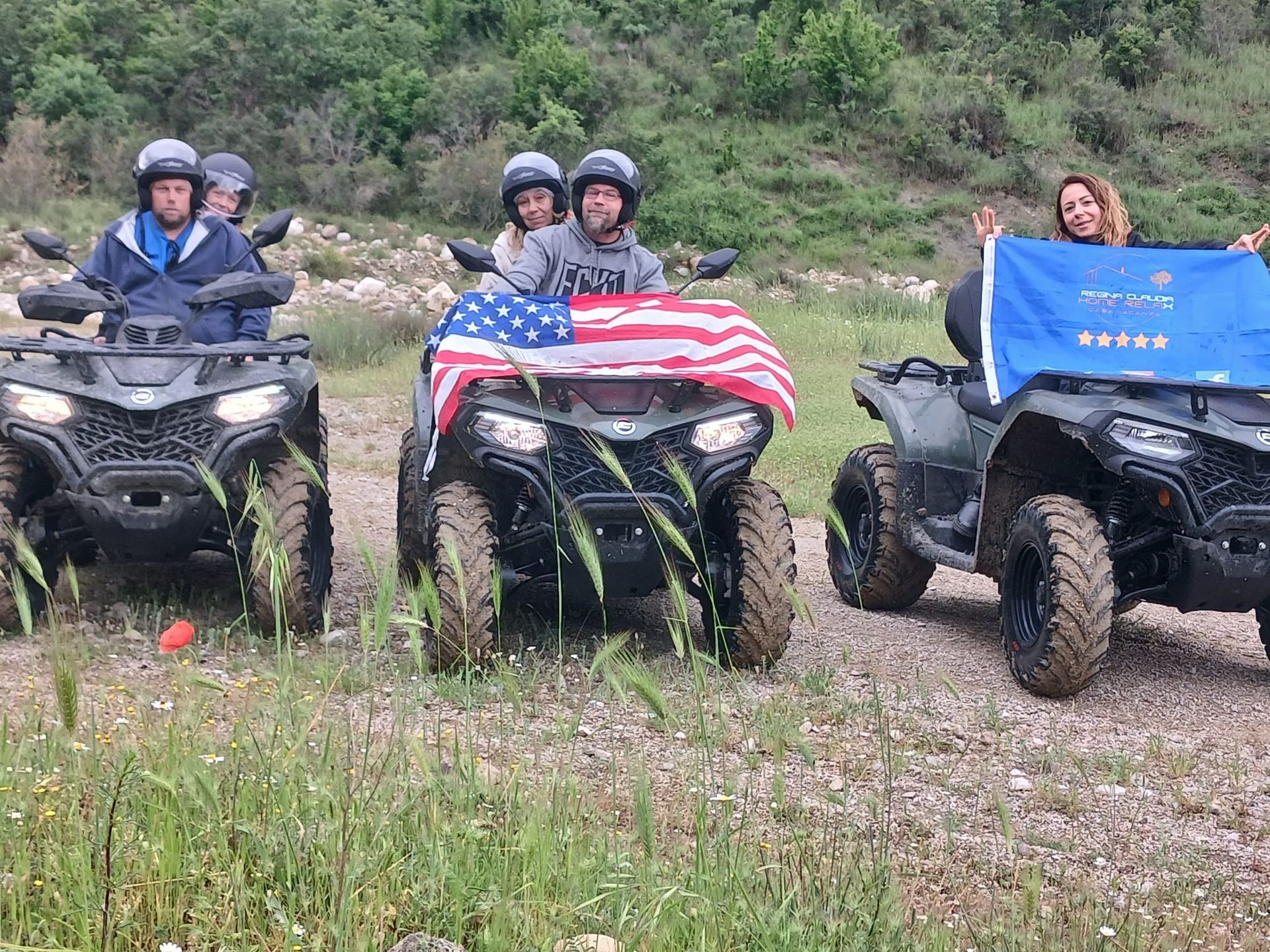 Three ATVs with riders on a rocky trail, one holding an American flag in front.