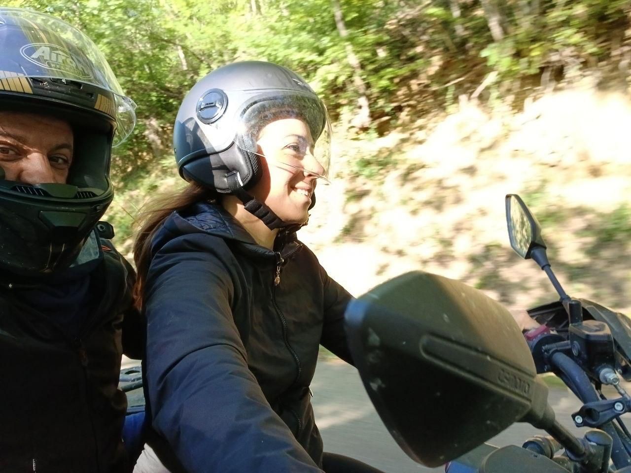 Two helmeted riders on a motorcycle, one smiling on a sunny wooded road.