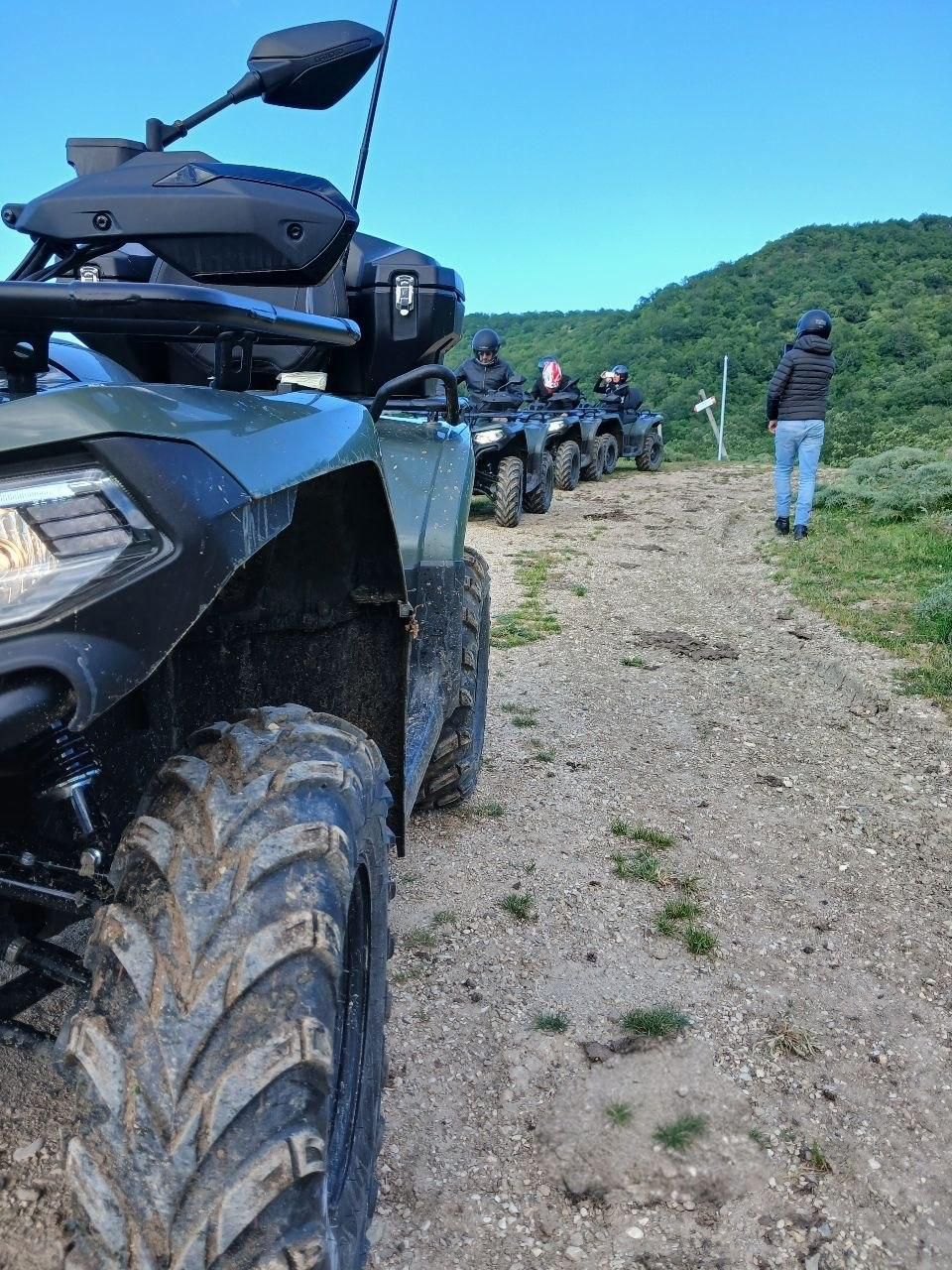 ATVs lined up on a dirt trail with riders in helmets near green hills under a blue sky