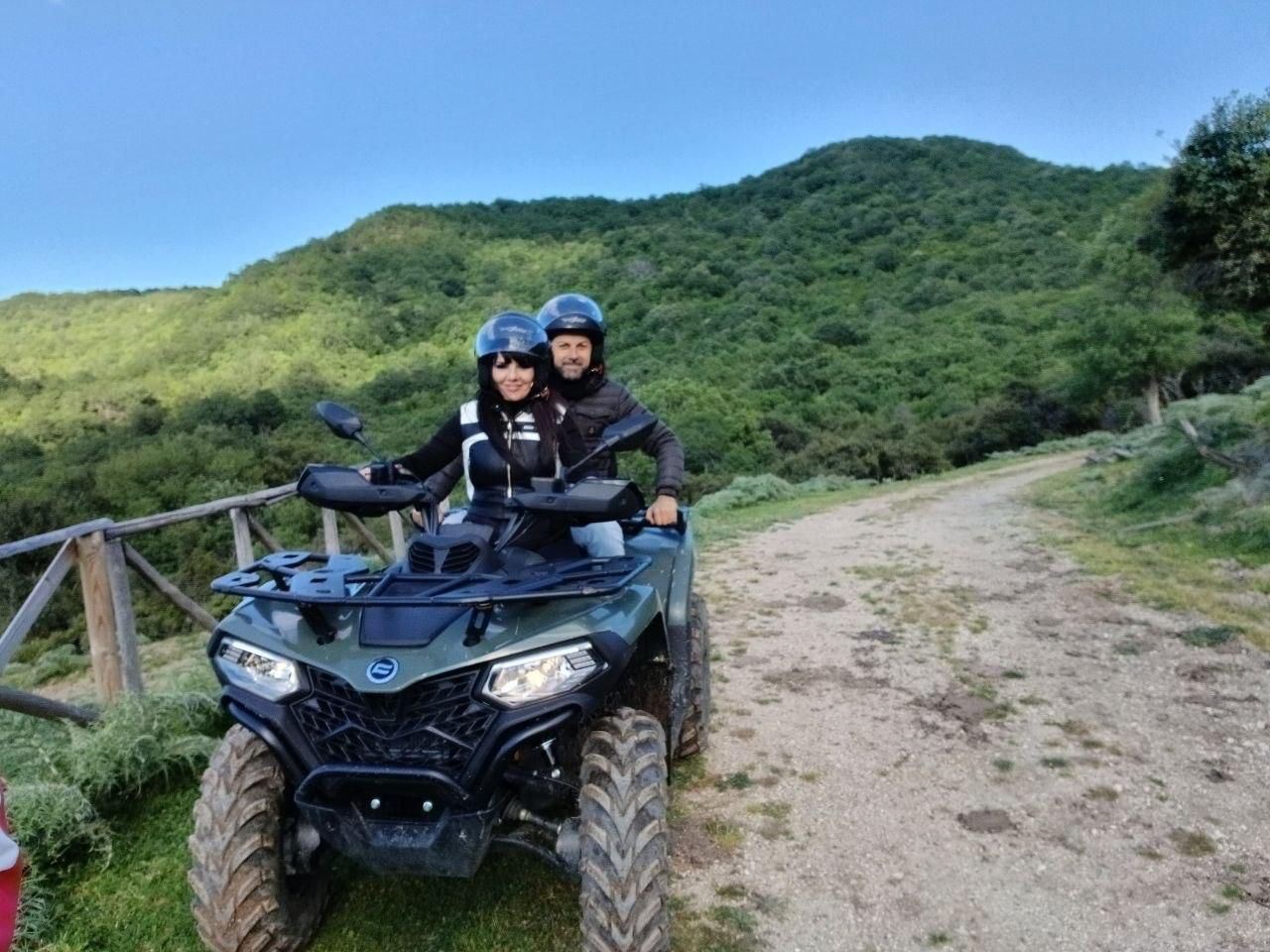 Two people on an ATV on a dirt trail with green hills in the background