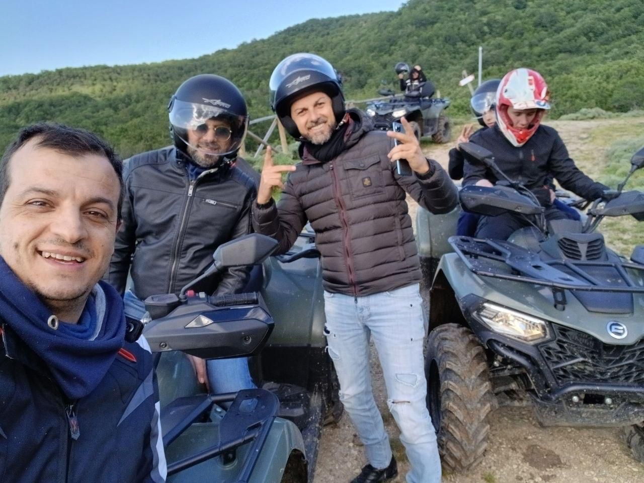 Four people on ATVs outdoors, smiling in helmets on a dirt trail with green hills behind them