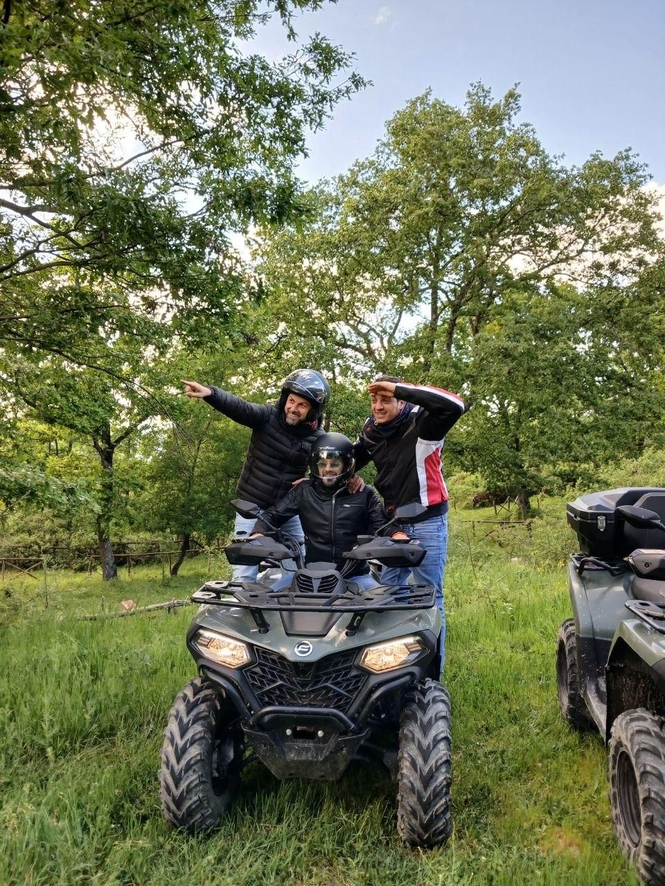 Three people posing on an ATV in a grassy wooded area