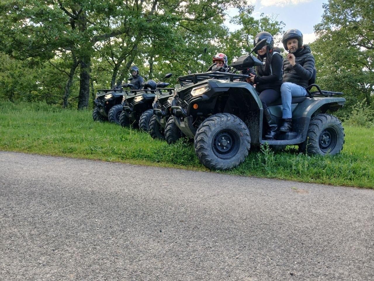 People riding ATVs on a grassy trail beside a road, with trees in the background
