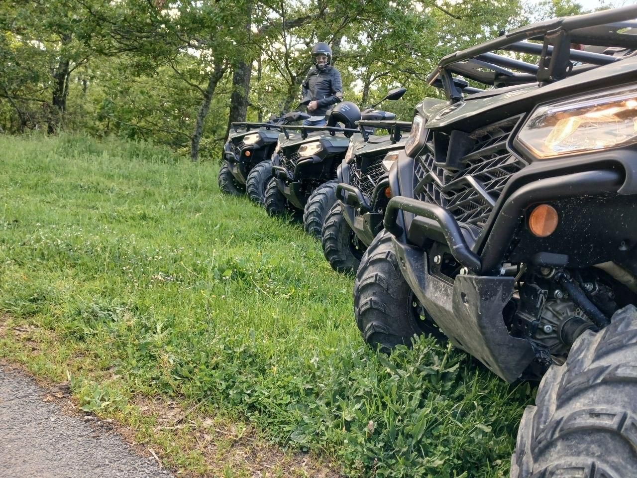ATVs lined up on a grassy trail edge, with a rider on the lead vehicle in a wooded area