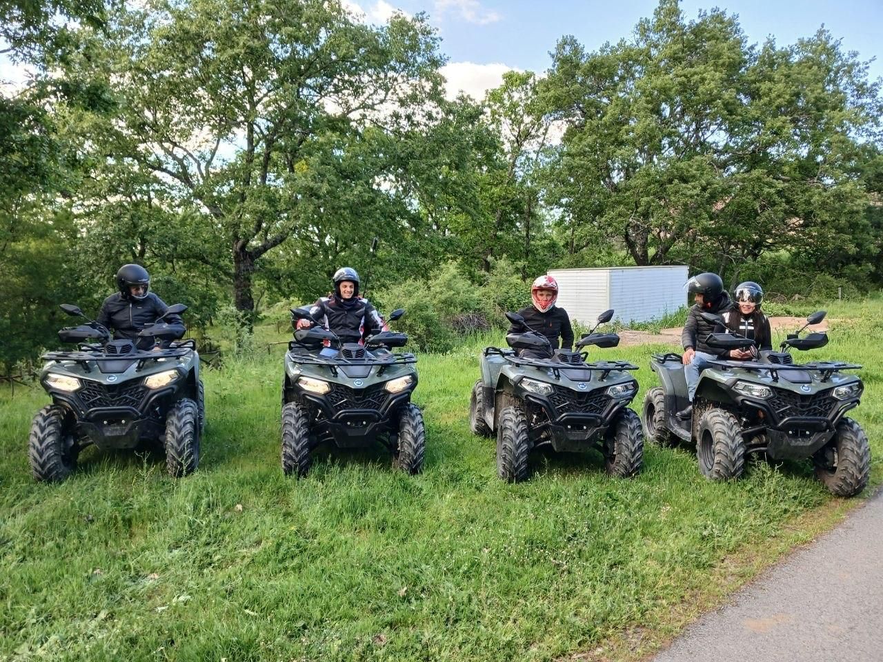 Four people on black ATVs lined up on a grassy trail beside trees and a road