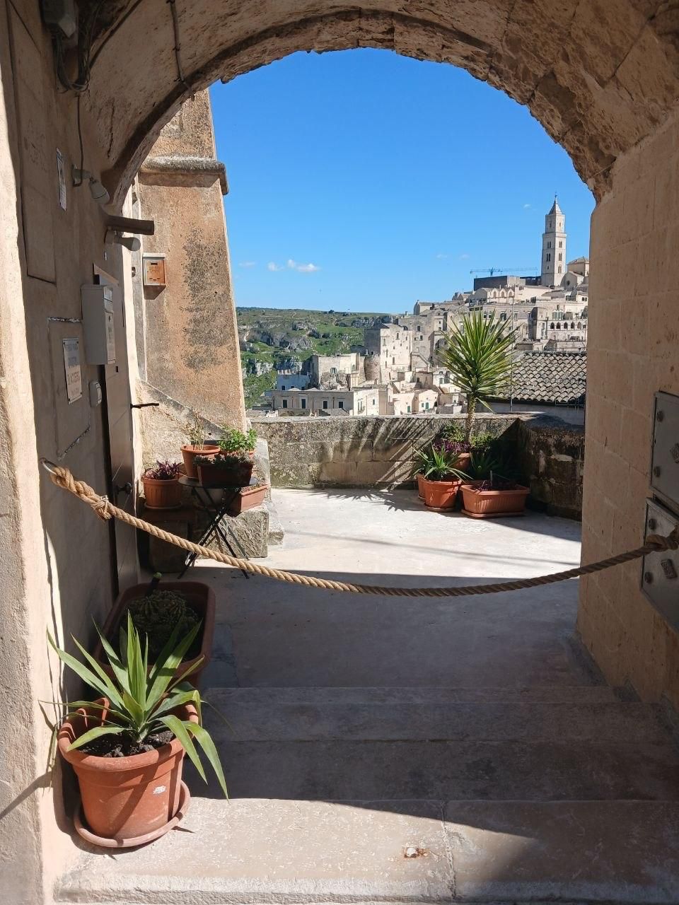 Stone archway framing a sunlit hilltop town with potted plants and a rope barrier in the foreground