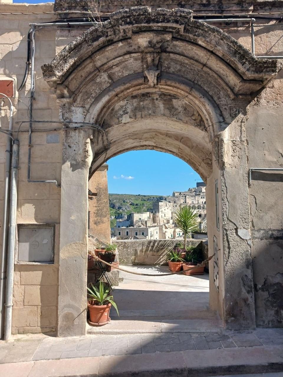 Stone archway opening to a sunlit courtyard and hillside view, with potted plants by the entrance