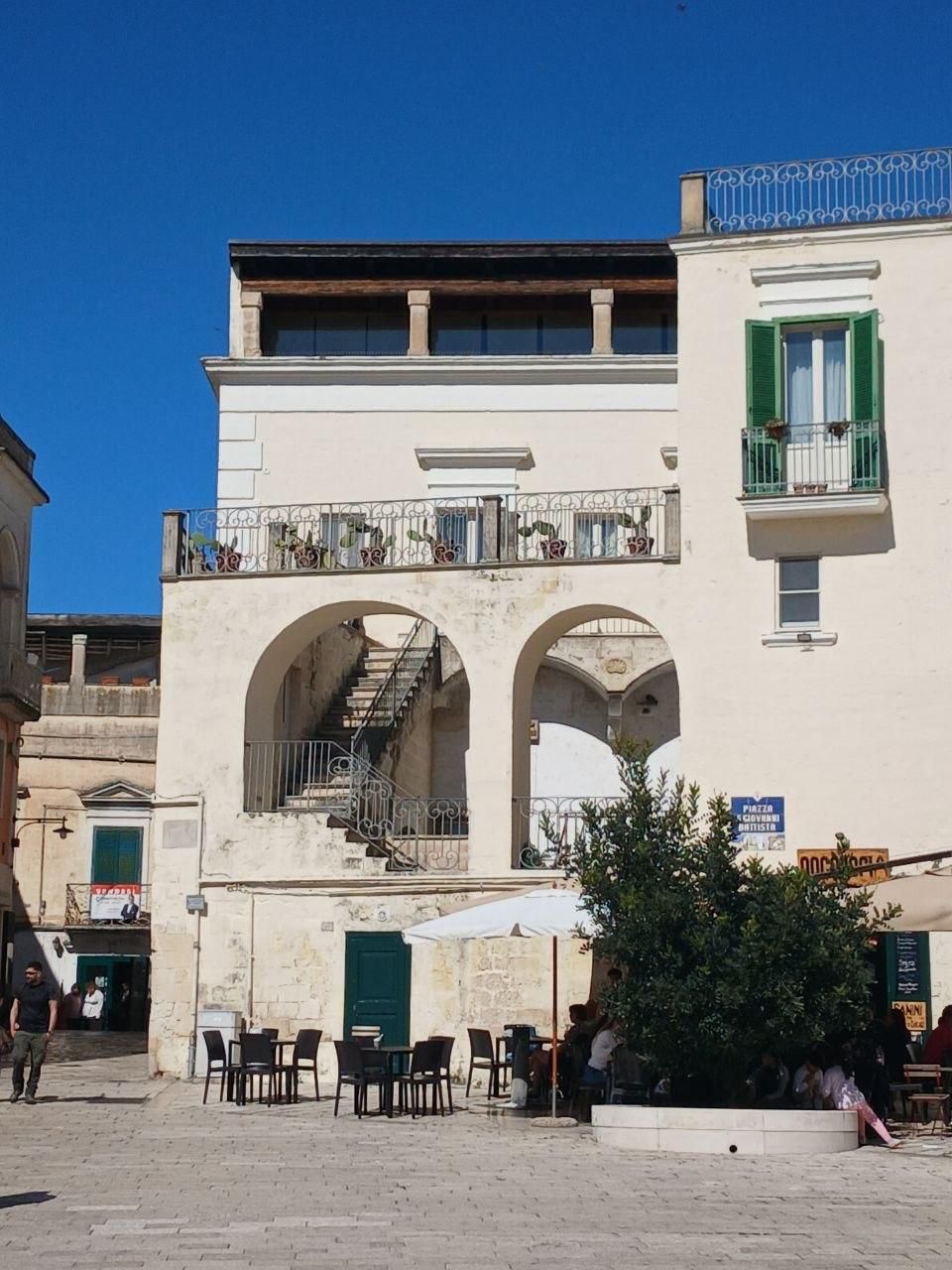 White stone building with arched openings, balcony, and outdoor café tables under a blue sky