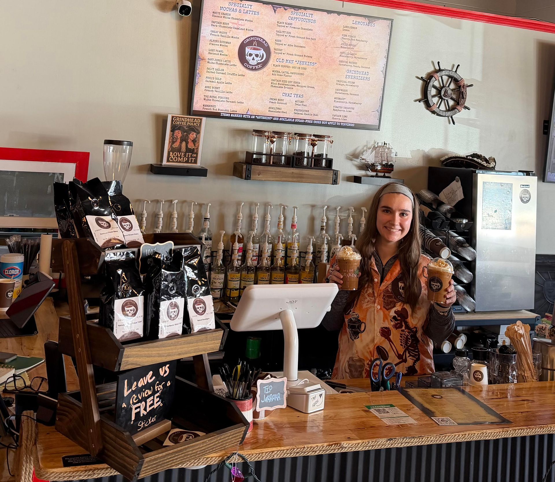 Woman behind a coffee shop counter holding iced coffees; wooden display with coffee bags.