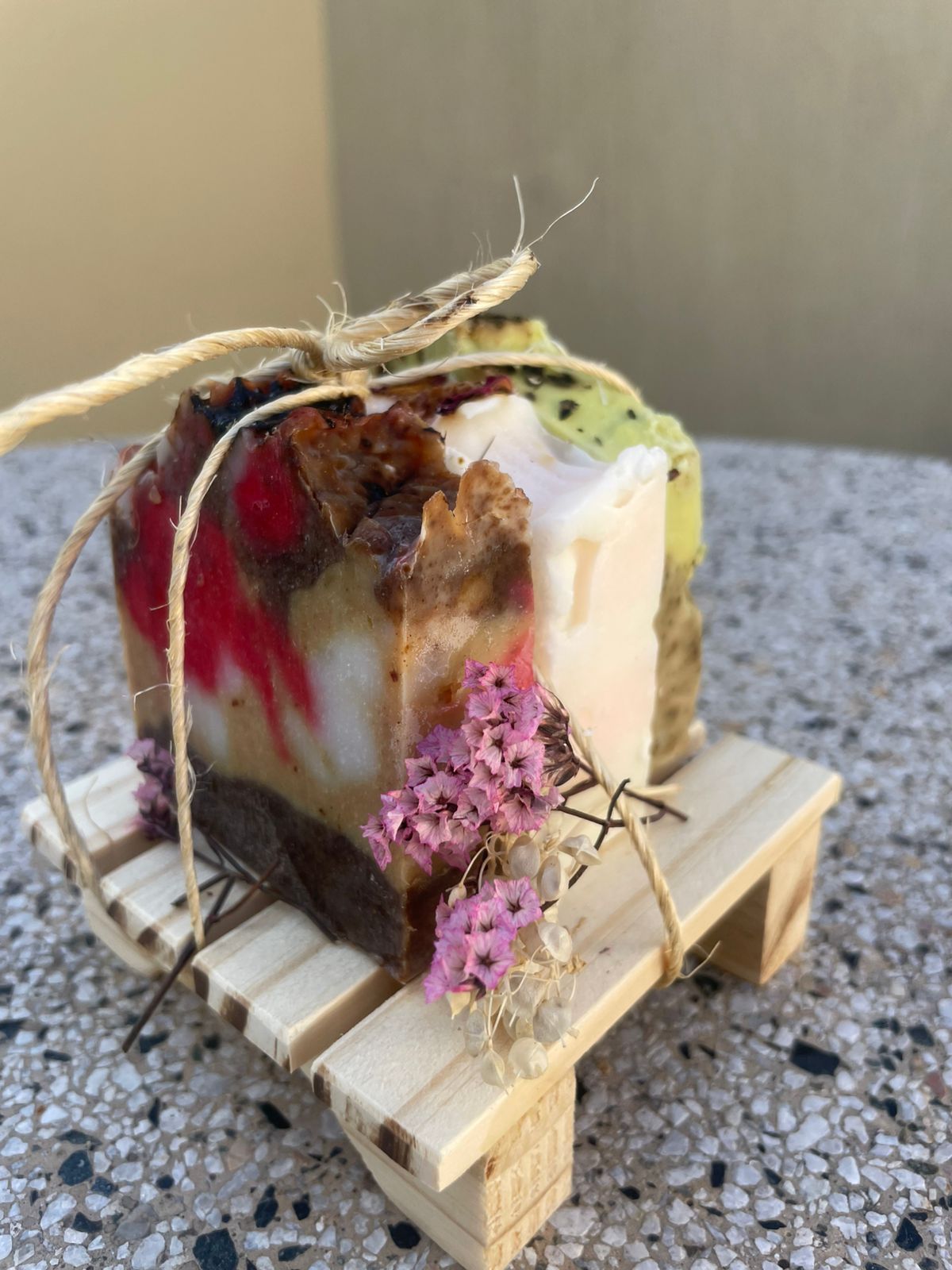 Barra de jabón colorida en capas atada con cordel sobre una pequeña paleta de madera, decorada con flores secas.