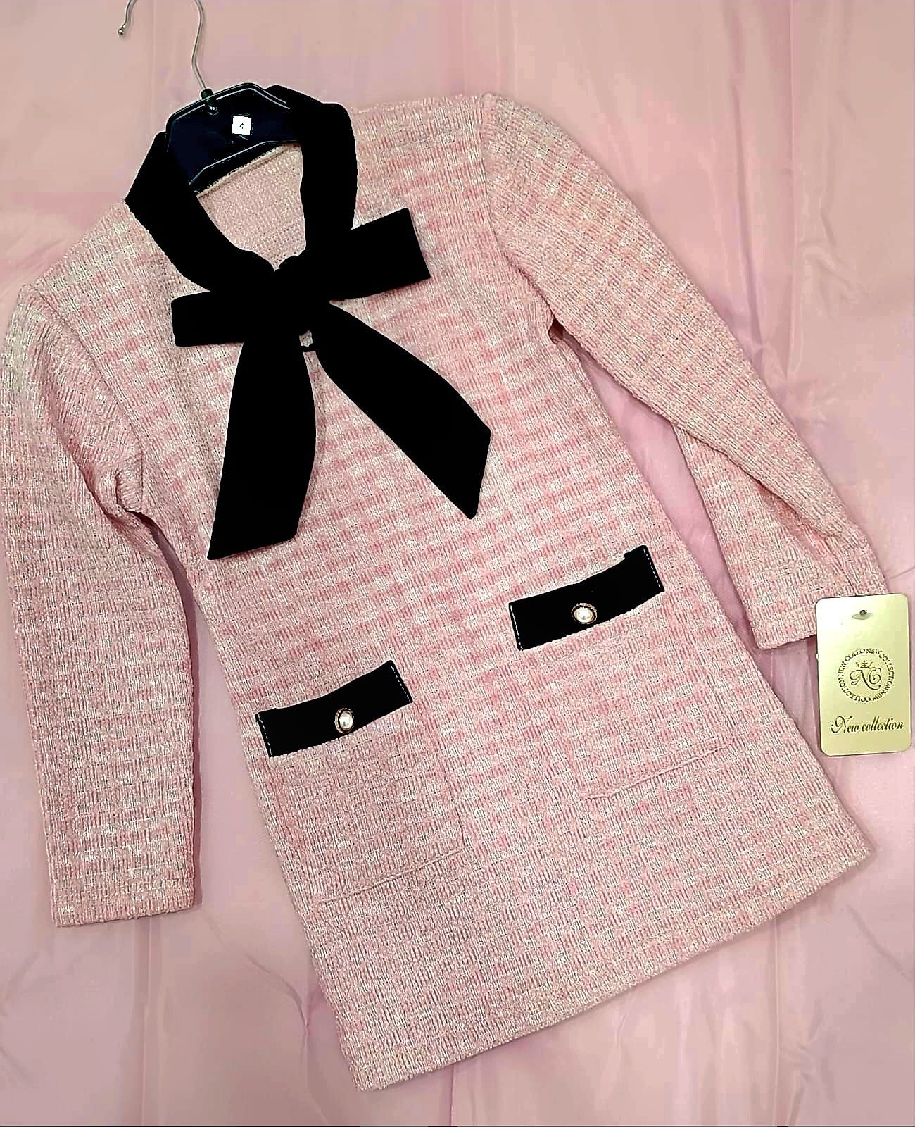Un vestito rosa con un fiocco nero e tasche è appeso a una gruccia.