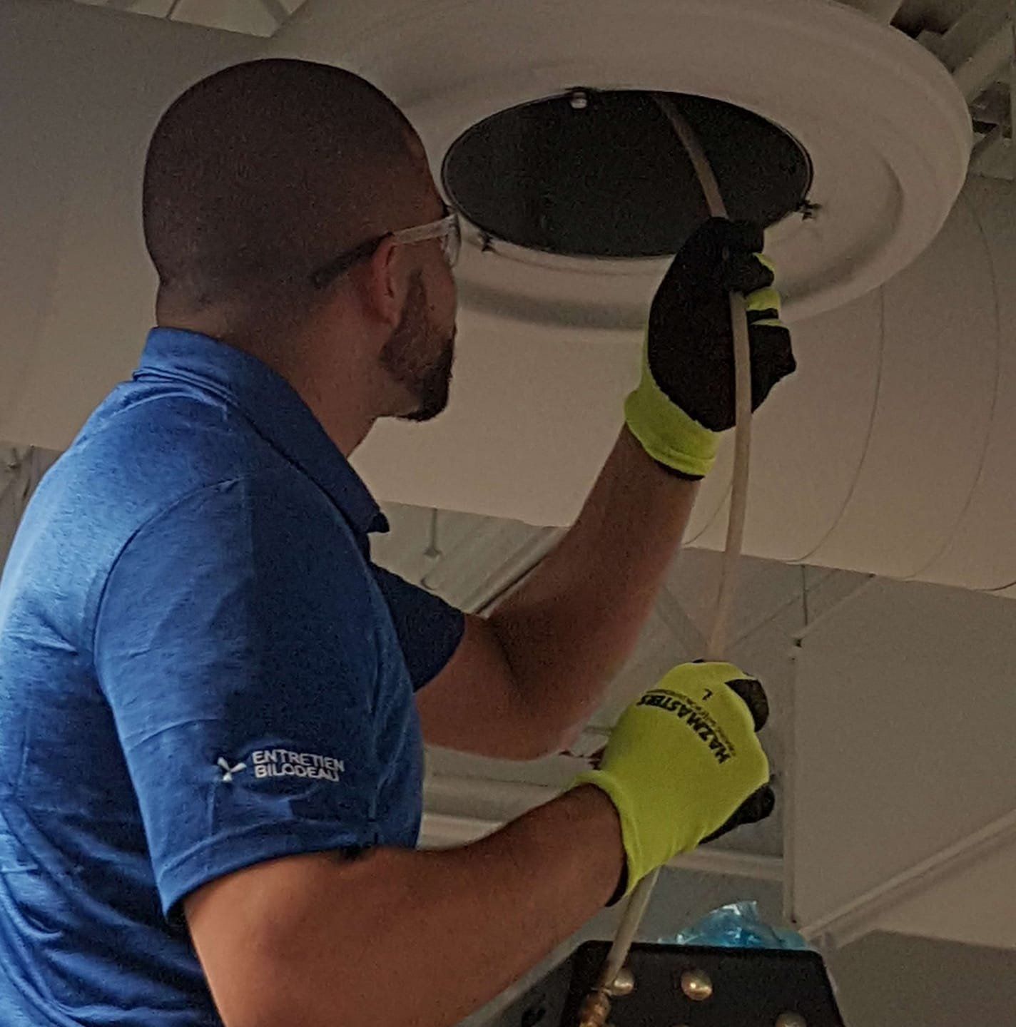 Un homme en chemise bleue et gants inspecte une grille d'aération, tenant un tube, dans un plafond blanc.
