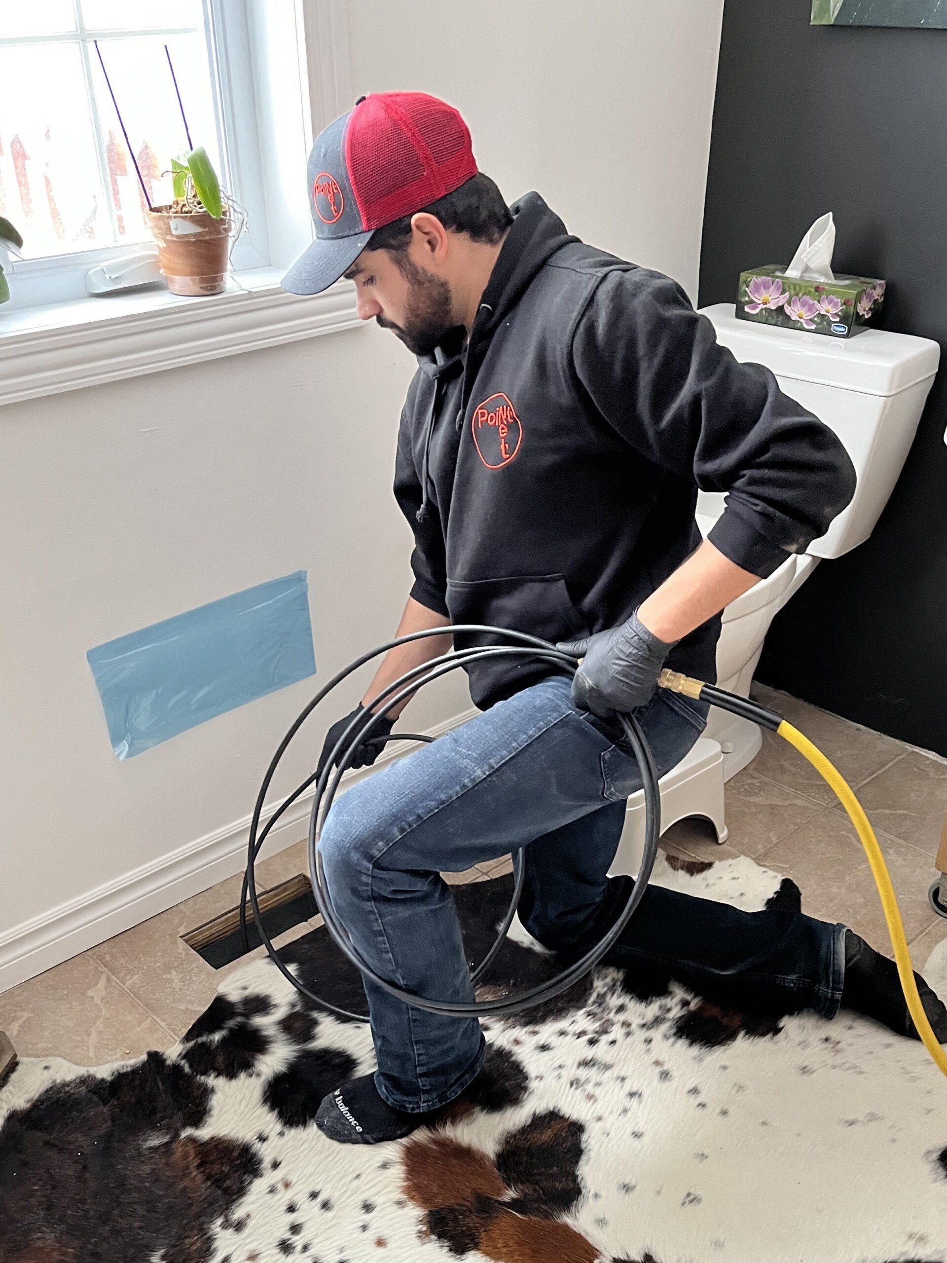 Un homme portant une casquette et des gants débouche une canalisation dans une salle de bains, à l'aide d'un cordon noir et d'un tapis en peau de vache.