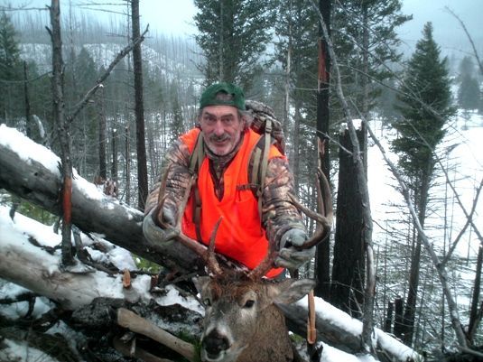 A man in an orange vest is standing next to a deer in the woods.